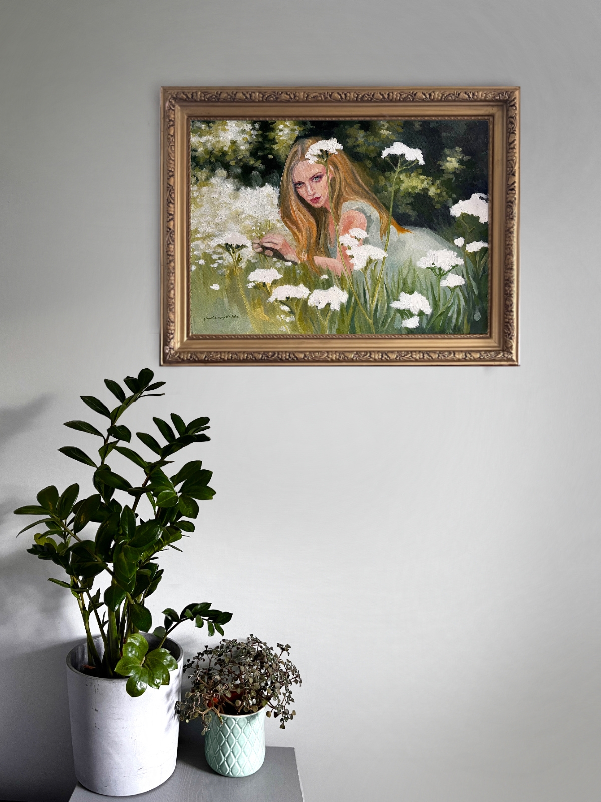 Vigil, original oil painting of a reclining woman in a flower meadow, shown in a gold frame on a light wall above potted plants.