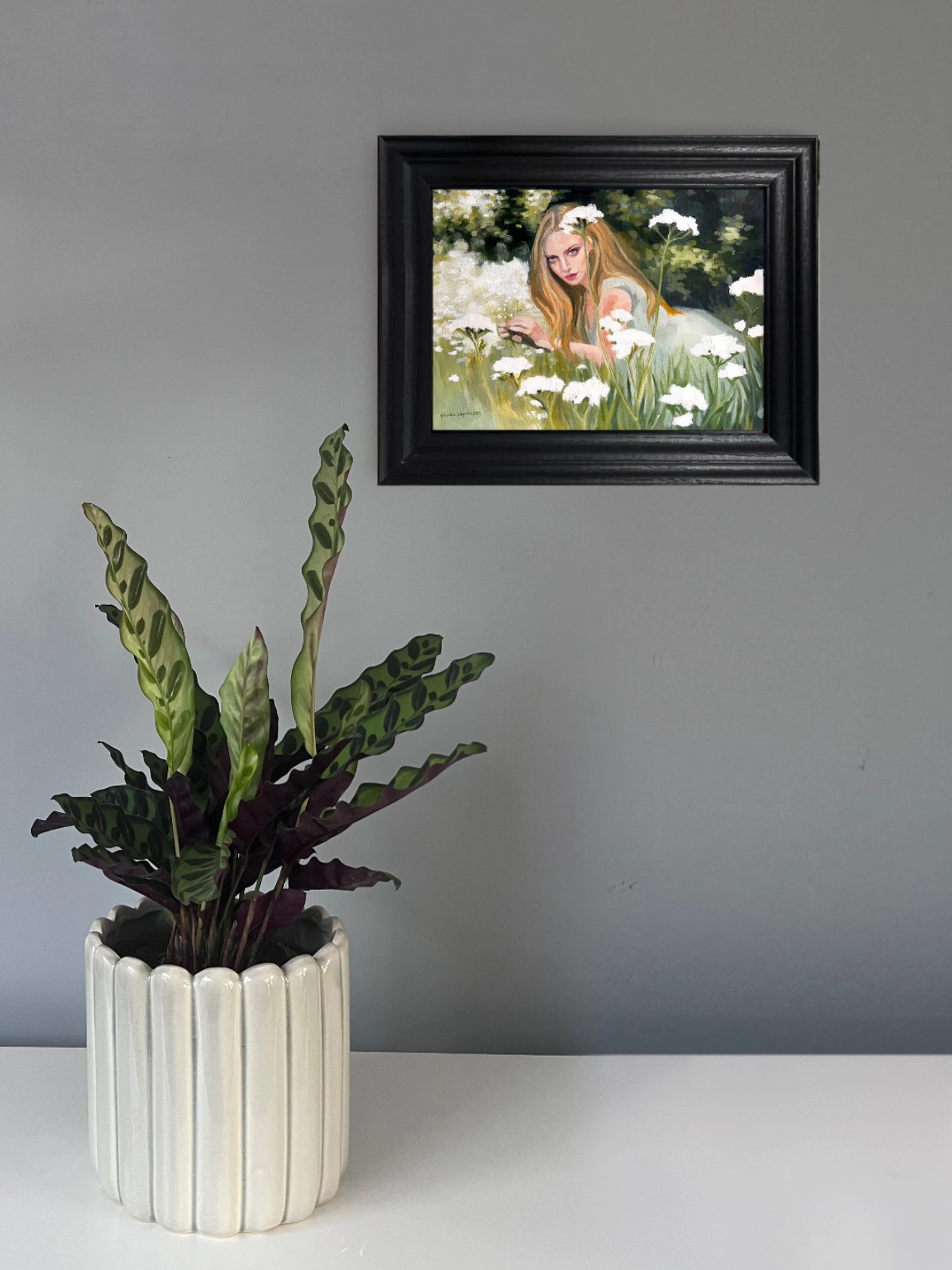 Blonde woman reclining among tall grasses and white wildflowers, looking directly at the viewer, with woodland behind her